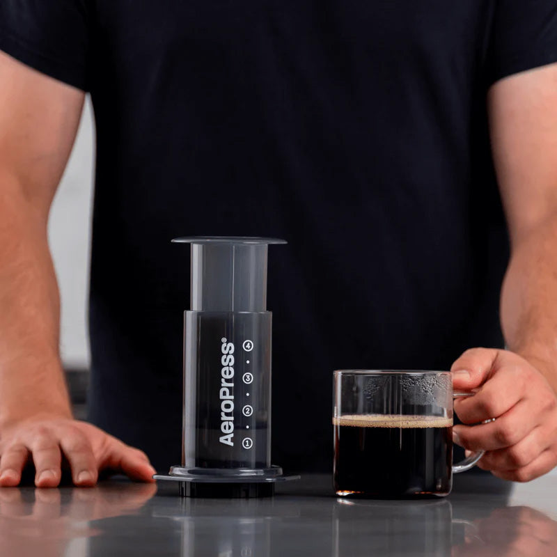 Assembled AeroPress Original on a counter beside a glass mug of brewed coffee; mug not included.