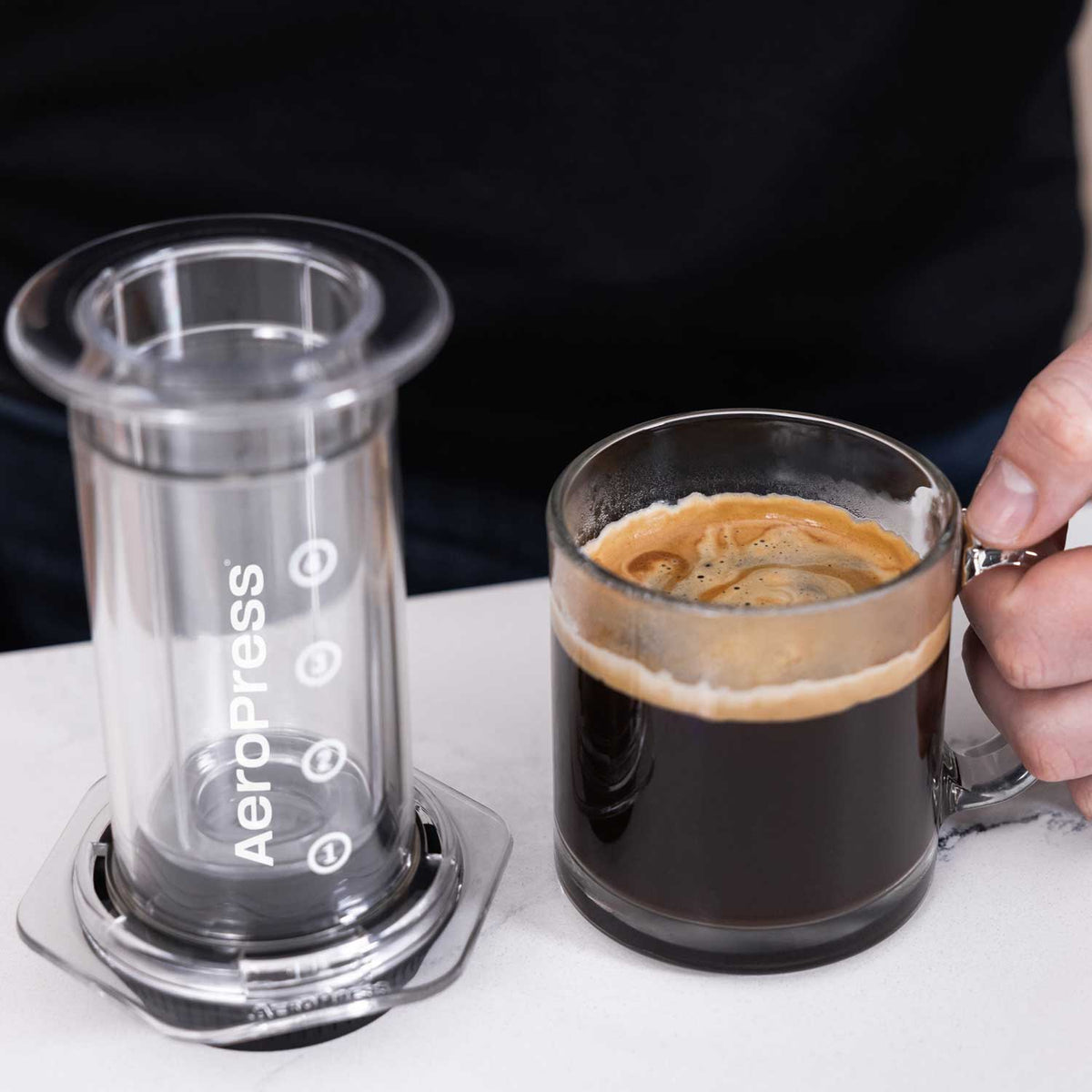 Clear AeroPress brewer fitted with the Flow Control Filter Cap beside a glass mug of coffee; brewer and mug not included.
