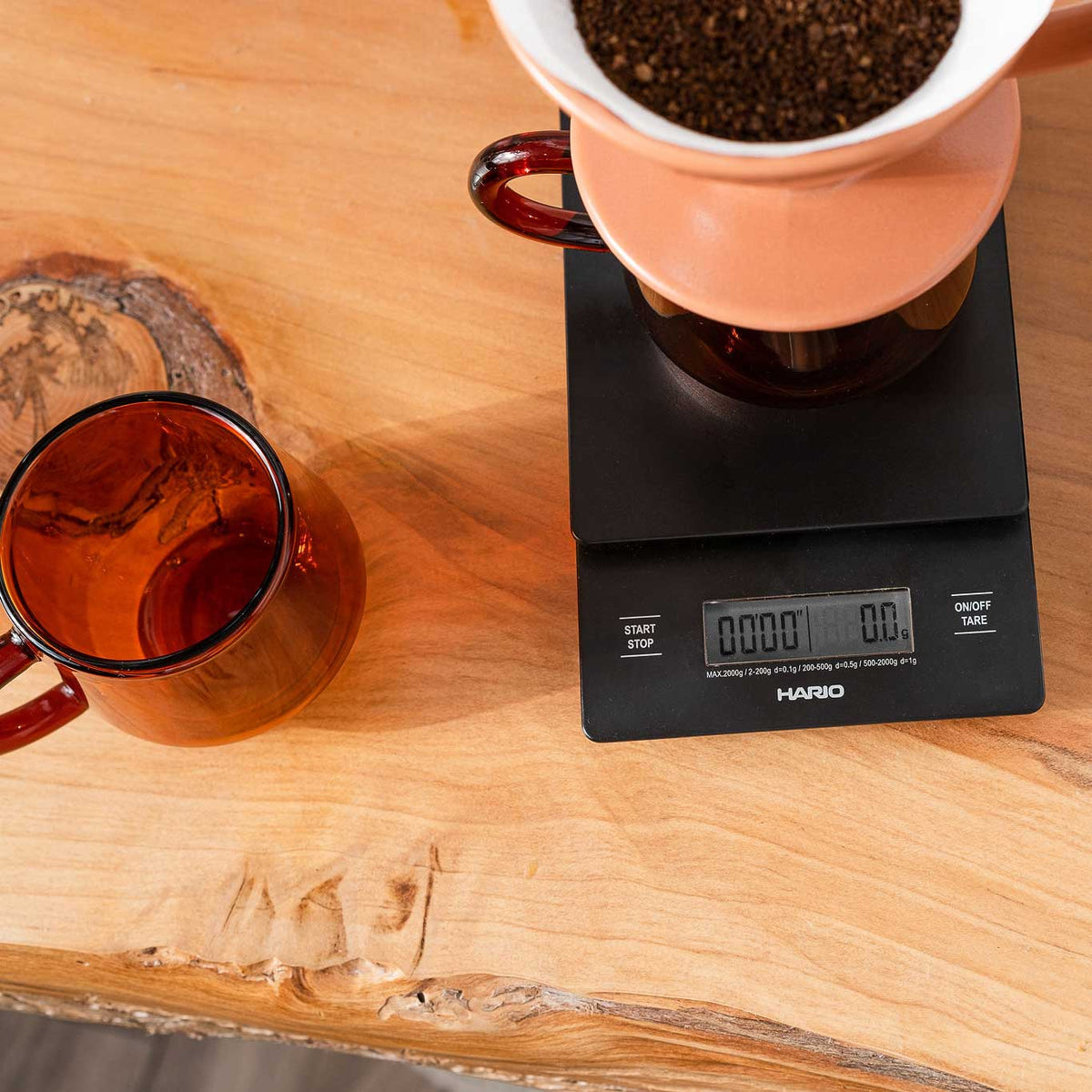 Hario V60 drip scale and timer on a wooden counter under a ceramic dripper, with a glass of coffee alongside; dripper and glass not included.