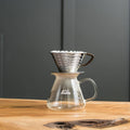 Kalita Wave 185 stainless steel dripper with a wave paper filter, resting on a glass Kalita server on a wooden table; server and filters not included.