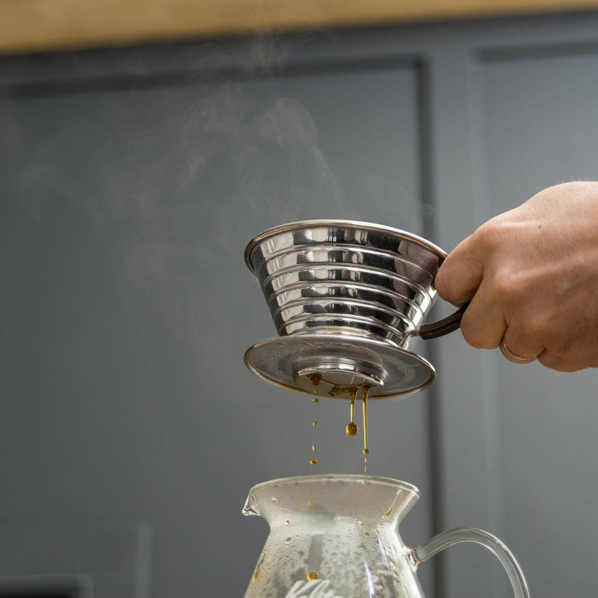 Hand lifting a Kalita Wave 185 dripper as brewed coffee drips into a glass server below; server not included.